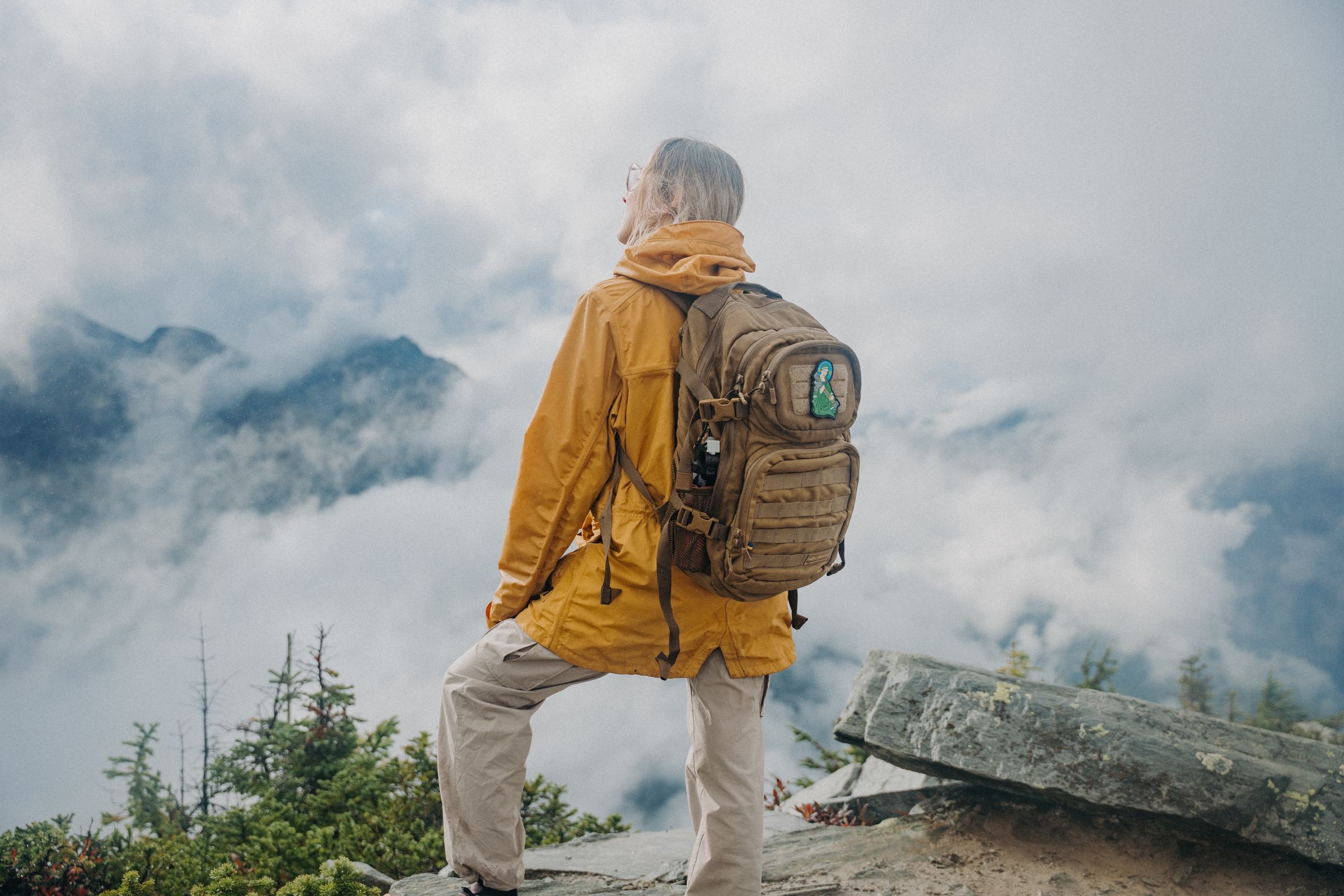 Hiker in a yellow jacket wearing a Saint Javelin backpack, looking out over a cloud-filled valley in the Carpathian Mountains, Ukraine