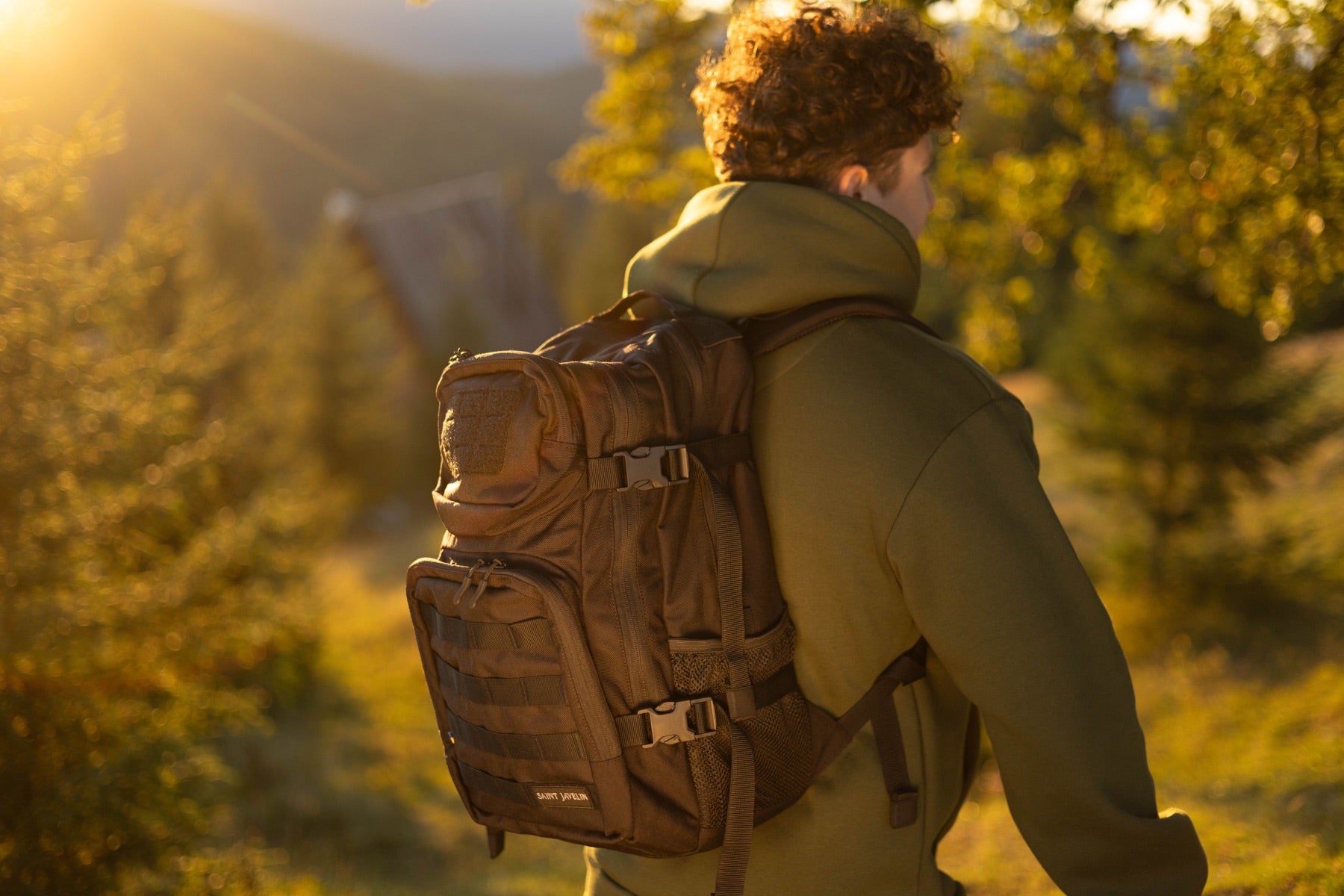Person seen from behind wearing a Saint Javelin backpack in golden light among pines, Carpathian Mountains, Ukraine